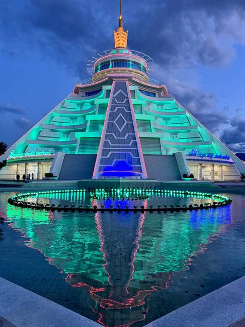       Futuristic turquoise-lit pyramid building reflected in a pool at dusk
  