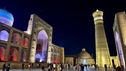       Night scene of Bukhara’s Po-i-Kalyan complex with the illuminated minaret and madrasah, visitors strolling below.
  