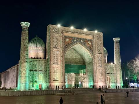       Sher-Dor Madrasah in Samarkand glowing with green and golden lighting against the night sky.
  