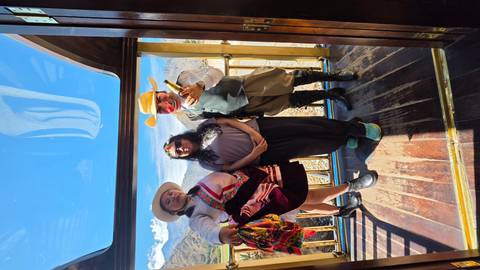       Three people, one in traditional costume, posing in the glass-roof observation car of an Andean train.
  