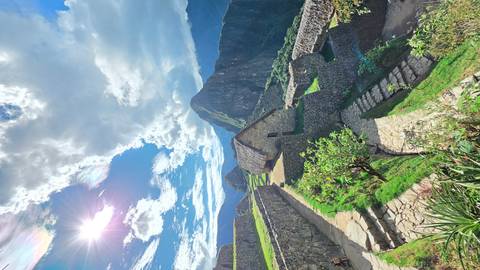       Sun-lit view of Machu Picchu terraces, stone houses and surrounding peaks beneath a dramatic sky.
  