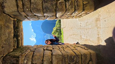       Tourist playfully peeking through a narrow stone doorway with Andes mountains beyond.
  