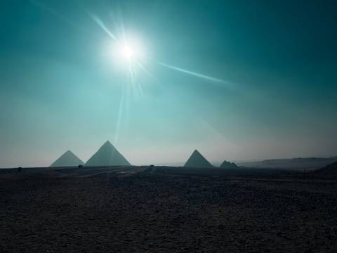       Silhouettes of the pyramids of Giza against a hazy desert horizon with a bright sun burst overhead.
  