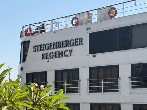       Close-up of the white river cruise ship façade with the name 'Steigenberger Regency' and life rings on the railing.
  