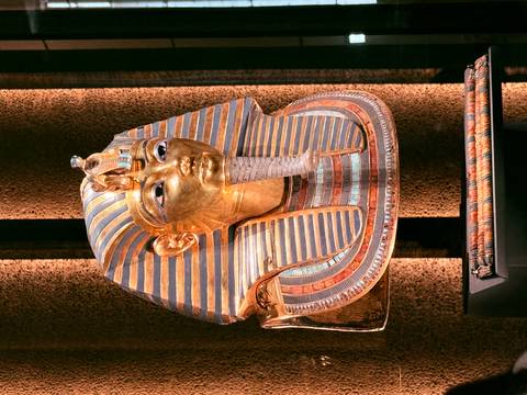       Gold and blue funerary mask of Tutankhamun displayed under spotlights against dark drapery.
  