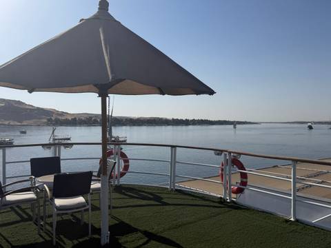      Open deck of a Nile cruise with umbrella-shaded table and panoramic river views on a bright day.
  