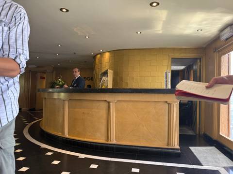       Hotel reception area with a gold-tiled front desk where a suited staff member greets arriving guests.
  