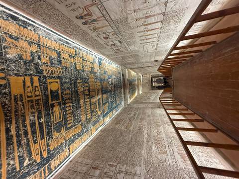       Fresco-lined corridor inside a pharaonic tomb with intricate painted ceiling leading visitors downward.
  