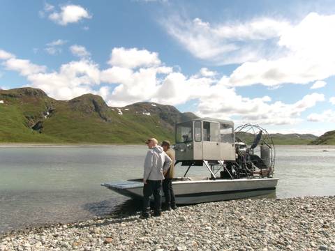       Two men stand on a shallow-draft boat beside a wide river backed by rolling green hills under scattered clouds.
  