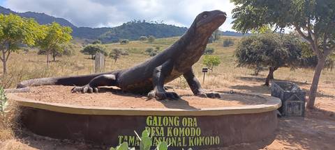       Large Komodo dragon statue on a plinth with signage in a dry grassy landscape.
  
