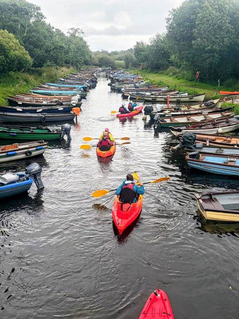       Three kayakers paddling through a marina channel lined with moored boats.
  