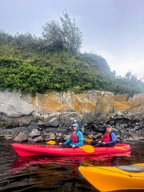       Two kayakers posing by a rocky, seaweed-covered shoreline beneath lush vegetation.
  