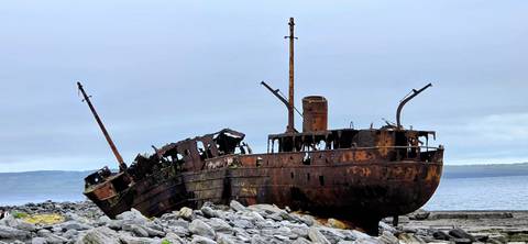       Rusty wreck of the Plassey ship stranded on rocky shoreline under a grey sky.
  