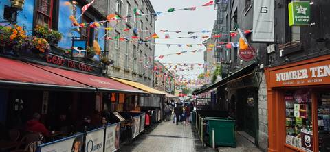       Colorful pedestrian street in Galway lined with pubs, bunting flags, and strolling visitors on a wet day.
  