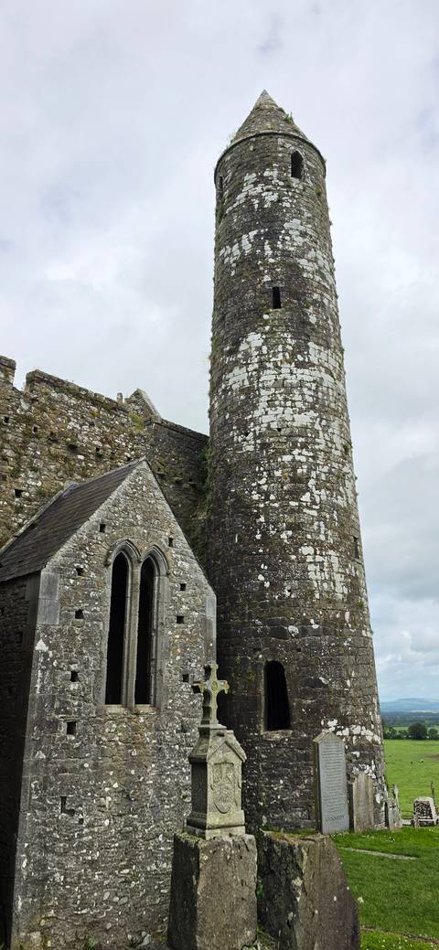       Ancient stone round tower and chapel ruins with weathered lichen under grey skies.
  