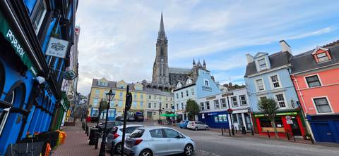       Colorful Georgian buildings lining a tidy street with St. Colman's Cathedral spire rising above in Cobh.
  