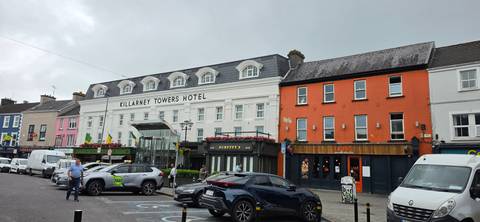       Streetscape of Killarney with Killarney Towers Hotel and shops on a grey day, cars parked in foreground.
  