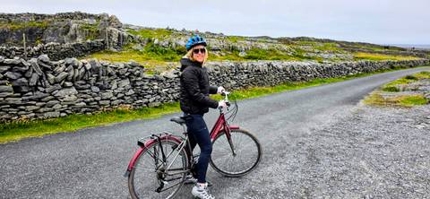       Smiling cyclist poses with red bike on rural lane bordered by stone walls and green fields
  