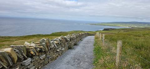       Gravel path lined by stone wall overlooking coastline and distant cliffs under cloudy sky
  