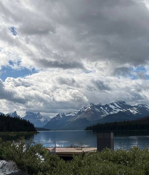       Snow-dusted Rocky Mountain peaks rise beneath dramatic clouds above a dark pine forest.
  