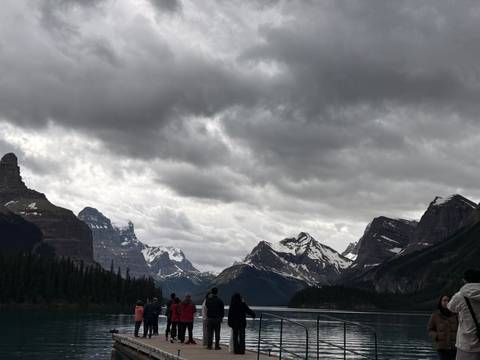       Tourists admire a glacial lake flanked by towering, brooding Rocky Mountains under heavy clouds.
  