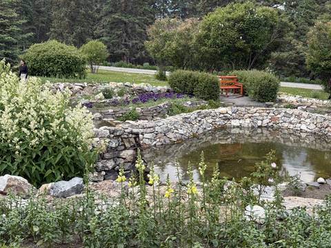      Stone-lined ornamental pond with blooming flowers and a red garden bench in a mountain park.
  