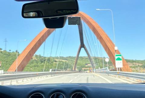       View through car windshield of modern rust-colored bridge arching over highway.
  