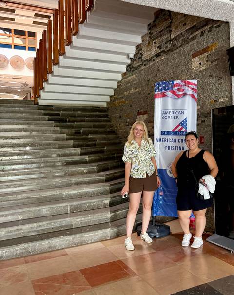       Two women pose by a banner that reads 'American Corner Pristina' inside a modern building lobby.
  