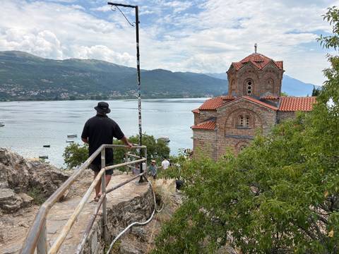       A traveler looks out over Lake Ohrid toward a cliff-top Byzantine church with mountains beyond.
  