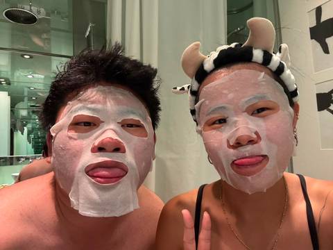       Two friends with sheet face masks stick their tongues out playfully in a bathroom
  