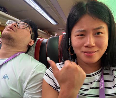       Woman takes a selfie pointing to her companion sleeping in a tour bus seat
  