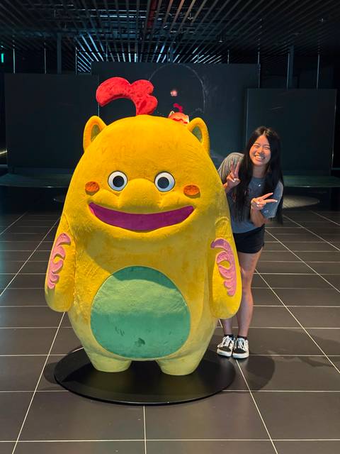       Traveller flashes peace signs next to a large cute yellow character statue in a modern hall
  