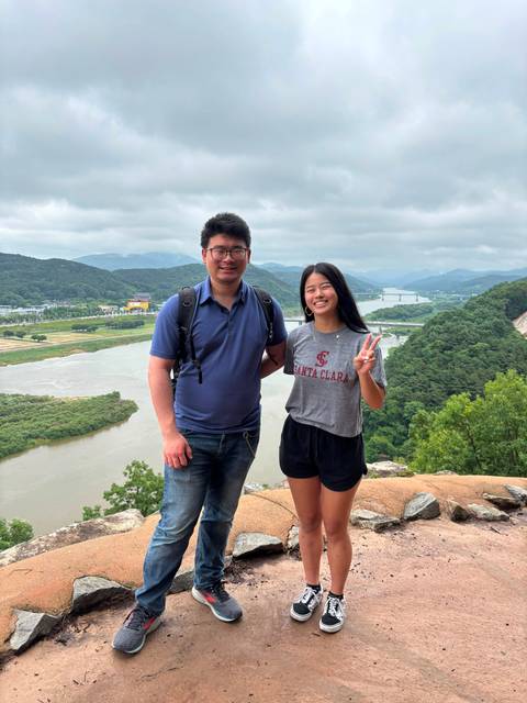       Couple stand on a cliff viewpoint overlooking a winding river and forested valley on a cloudy day
  