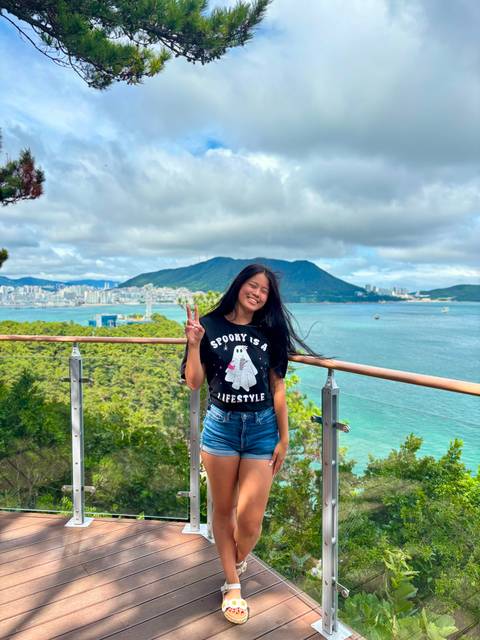       Woman smiles on a wooden deck overlooking turquoise sea and city skyline with forested hills
  
