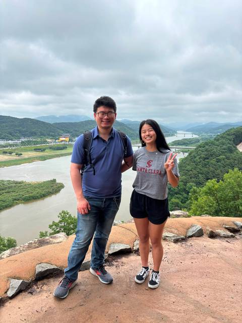       Same couple at the riverside lookout smiling with peace sign against valley backdrop
  
