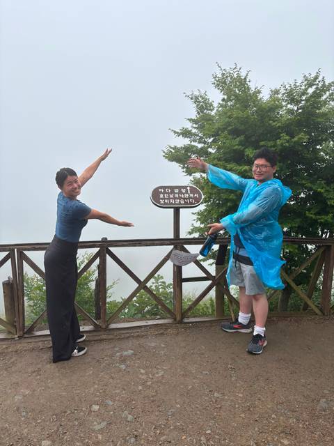       Two travellers in rain ponchos pose with arms outstretched on a fog-shrouded mountain deck
  