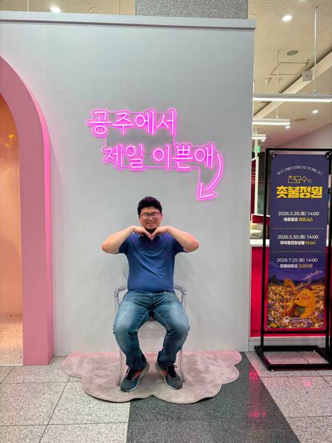       Man sits on transparent chair making a heart gesture under a pink neon Korean sign backdrop
  
