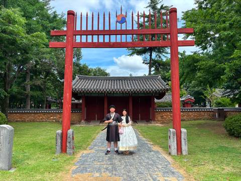      Couple dressed in traditional hanbok stand under a red wooden gate at a historic Korean complex
  