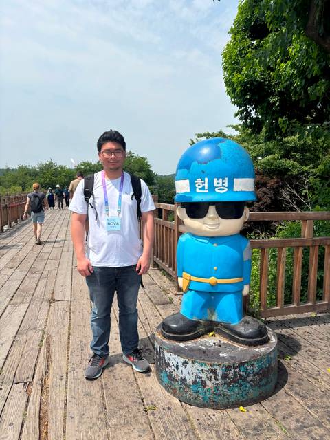       Traveller poses beside a blue cartoon soldier statue on a wooden bridge in sunny weather
  