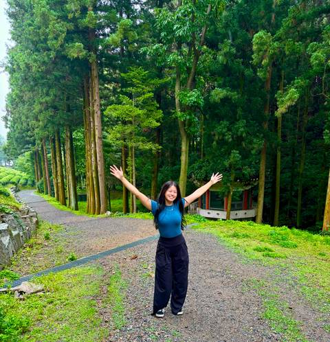       Woman raises her arms happily on a path lined with tall cedar trees in a verdant tea region
  
