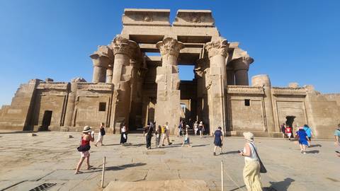       Visitors exploring the grand sandstone entrance of Kom Ombo Temple under clear blue sky
  
