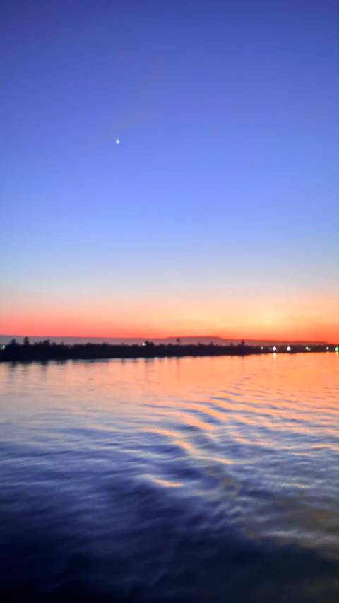       Blurry sunset fades from orange to deep blue over a calm river with faint lights on the horizon.
  