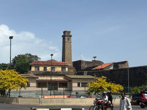       Historic clock tower rising above fort walls with flowering trees and clear blue sky.
  