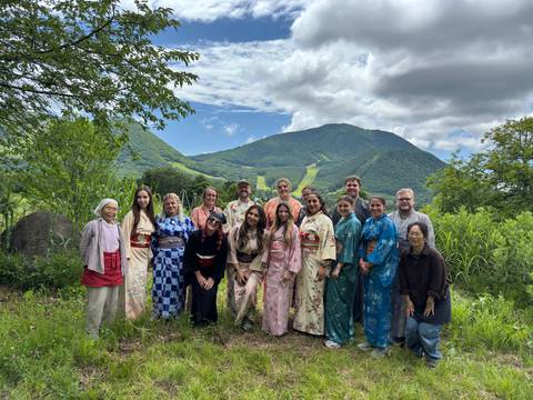       Large tour group wearing colourful kimonos pose in front of green mountain slopes under a blue sky.
  