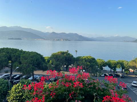       Scenic view over a calm lake framed by pink flowers, tree-lined shore and hazy mountain backdrop under clear blue sky.
  