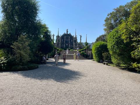       Visitors stroll a gravel path toward an elaborate baroque garden monument surrounded by lush manicured greenery on a sunny day.
  