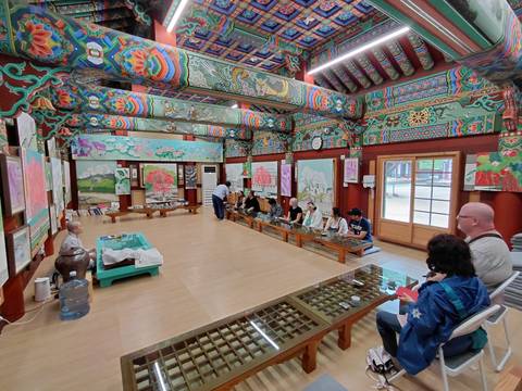       Visitors seated on the floor listening to a monk inside a vibrantly painted Korean temple hall
  