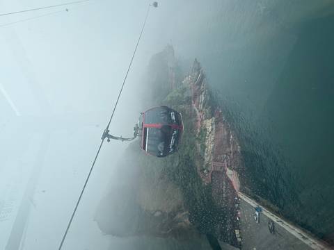       Enclosed red cable car travelling over a misty coastline with low visibility
  
