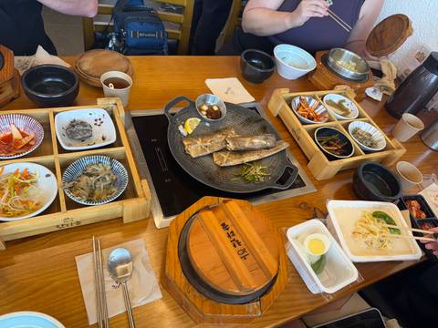       Korean dining table setup with grilled fish on a hot plate and several colorful side dishes presented neatly on wooden trays.
  
