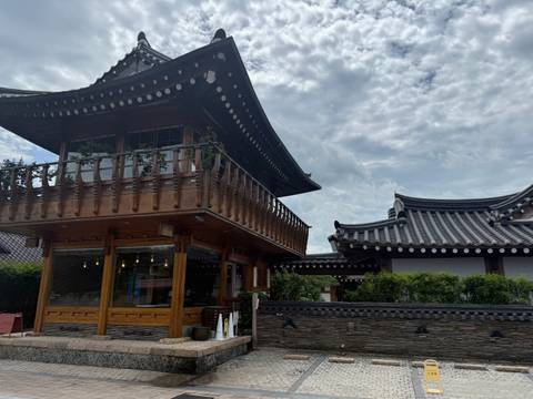      Wooden two-storey hanok building and tiled roof structures under a partly cloudy sky.
  